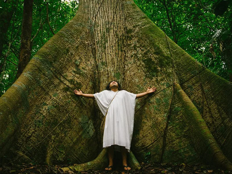 Documentary scene of a person in a white traditional tunic with arms open before a giant ceiba tree in the jungle